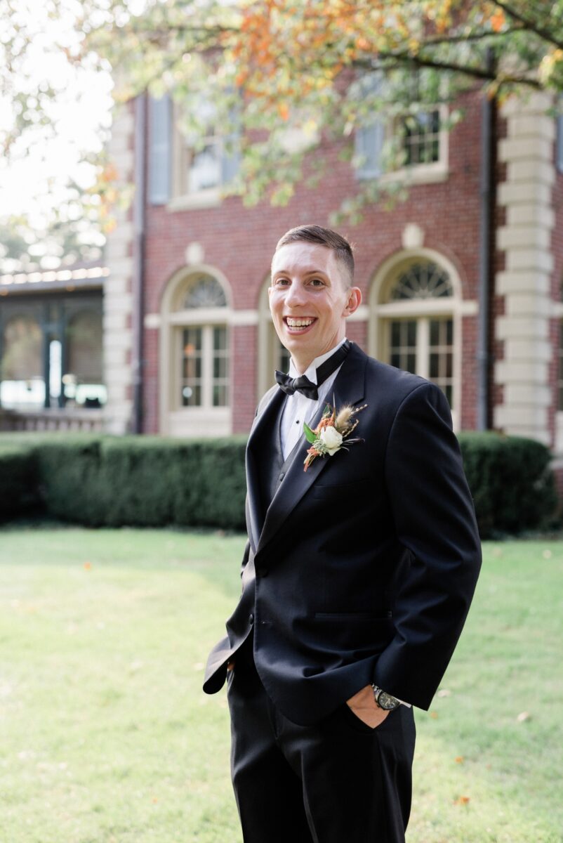 Portrait of the groom with black tux and coral and white boutonniere  smiling at the camera with the mansion on the background.