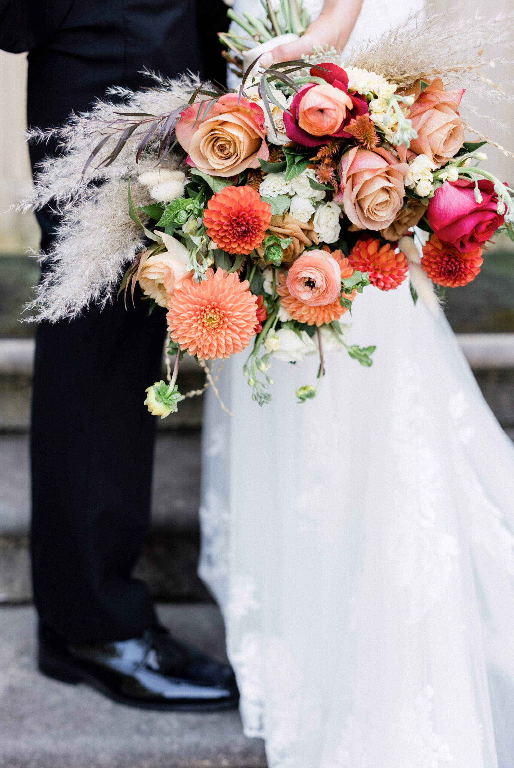Closeup of bridal bouquet with roses, dahlias and ranunculus in shades of coral and pink framed with pampas grass with the bride and groom in the background photographed by Maria Silva Goyo Photography