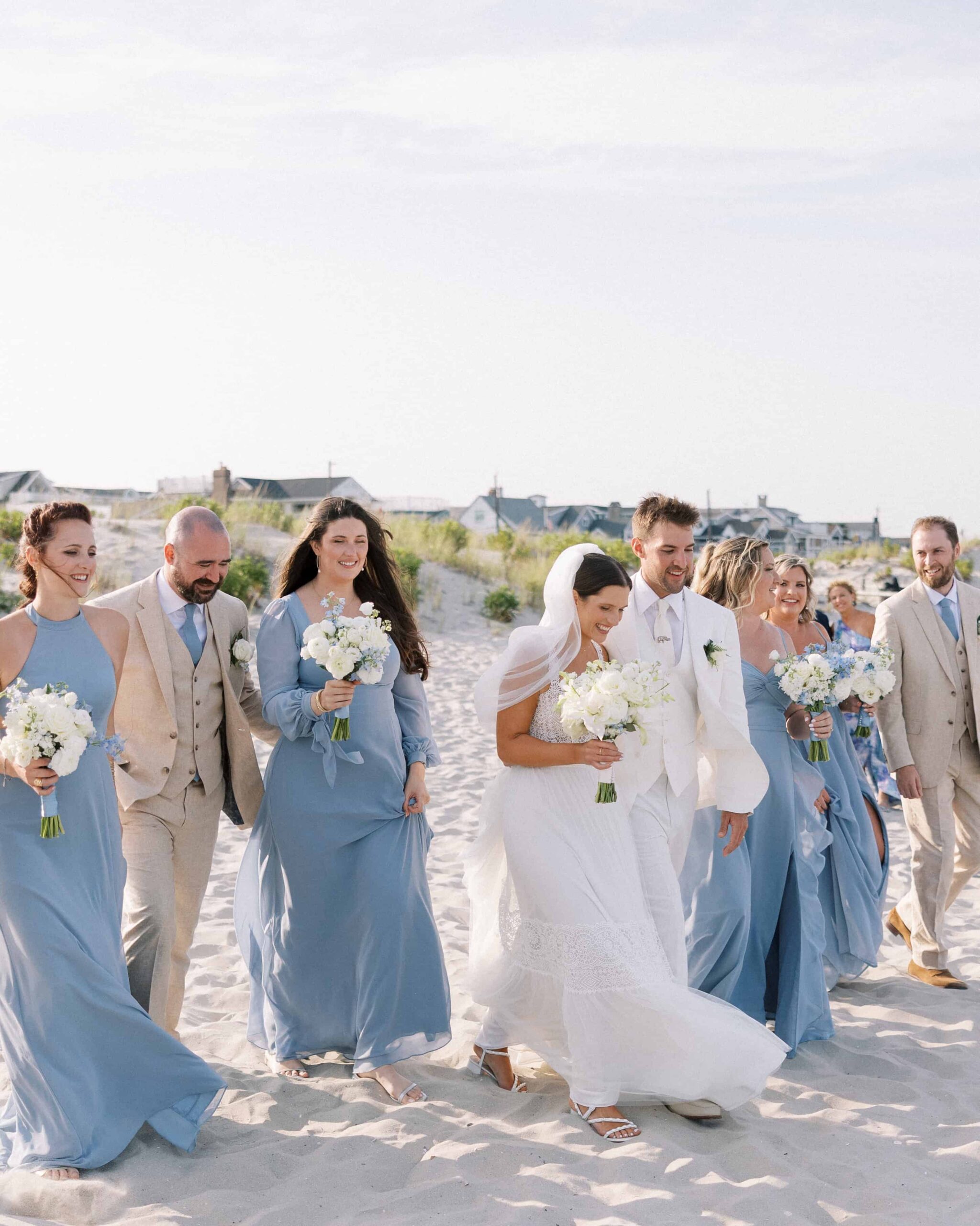 Bride and groom in white, walking on the beach with the wedding party wearing dusty blue dresses and beige suits.